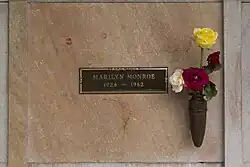 Photo of Monroe’s crypt, taken in 2005. «Marilyn Monroe, 1926—1962» is written on a plaque. The crypt has some lipstick prints left by visitors and flowers are placed in a vase attached to it.