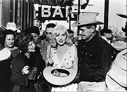 Monroe holding a hat and standing in the middle of a crowd of people, facing the camera. On her right is Gable and on her left, Winwood. There is a sign that says 'BAR' in the background.